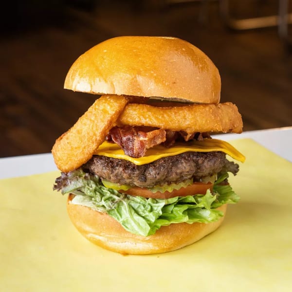 A tall burger with cheese, bacon, onion rings, lettuce, tomato, and pickles on a bun.